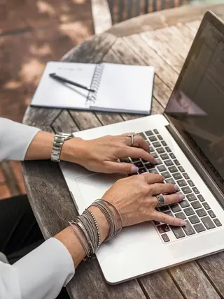 Eine Person sitzt im freien an einem Holztisch vor dem Computer und verfasst einen Text. Links neben dem Computer liegt ein Notizbuch und ein Stift.