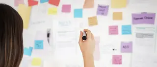 Eine Person steht vor einer Wand an die viele Post-Its geklebt wurden. In der rechten Hand hält sie einen Stift.