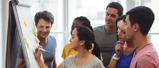 5 Personen stehen in einem hellen Raum gesammelt vor einem Flipchart. Eine Person in der Bildmitte schreibt mit einem Stift etwas auf das Flipchart.